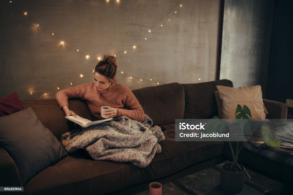 Woman reading book in cozy living room Young woman reading book and drinking coffee on sofa in hygge house. Caucasian female relaxing in cozy living room and reading a book. Blanket Stock Photo