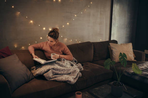Woman reading book in cozy living room Young woman reading book and drinking coffee on sofa in hygge house. Caucasian female relaxing in cozy living room and reading a book. hygge photos stock pictures, royalty-free photos & images