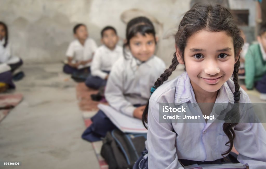 School girl in uniform of Indian Ethnicity sitting in their village classroom, looking at camera smiling. Girl sitting on the floor of her government primary school in uniform along with some of her class mates sitting behind her.  Selective focus, shallow depth of field. India Stock Photo
