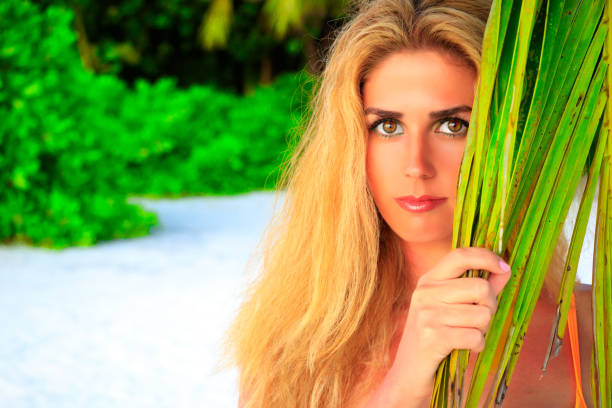 Beautiful blonde woman looks between palm leaves on the sandy beach at the camera Beautiful blonde woman looks between palm leaves on the sandy beach at the camera meeru island stock pictures, royalty-free photos & images