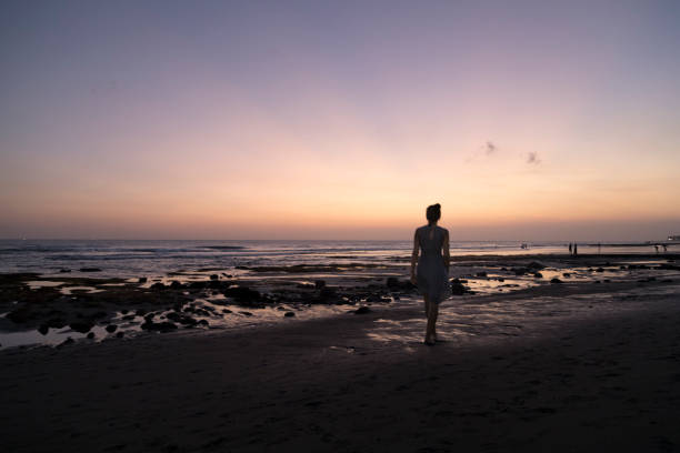 A woman in a dress walks along the tideline of a beach at sunset.  She is facing away from the camera, has her hair in a bun on the top of her head, has olive skin, and is walking along the ocean.  Shot at dusk on Canggu beach in Bali, Indonesia. Some motion blur in the figure.