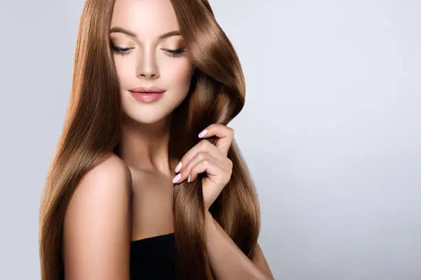 Young, brown haired woman  with voluminous hair.Beautiful model with long, dense, straight hairstyle and vivid makeup, is touching own hair with tenderness. Symbol of attentiveness to hair and good care of it. Incredibly dense, wavy,and shiny hair.