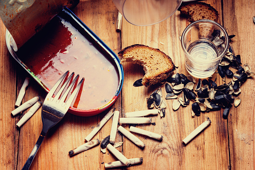 Photography of a open empty canned fish,fork,shots, seeds husk, bitten bread and cigarette butt