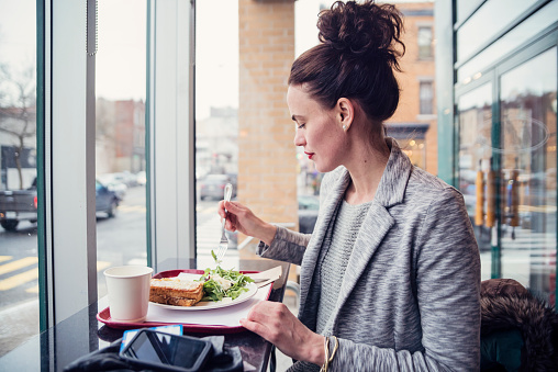 This is part of a series of the same woman in daily life, from morning jog to getting at work in winter. Here she is eating lunch in a city cafe. Horizontal waist up shot from indoors with copy space. This was taken in Montreal, Canada.
