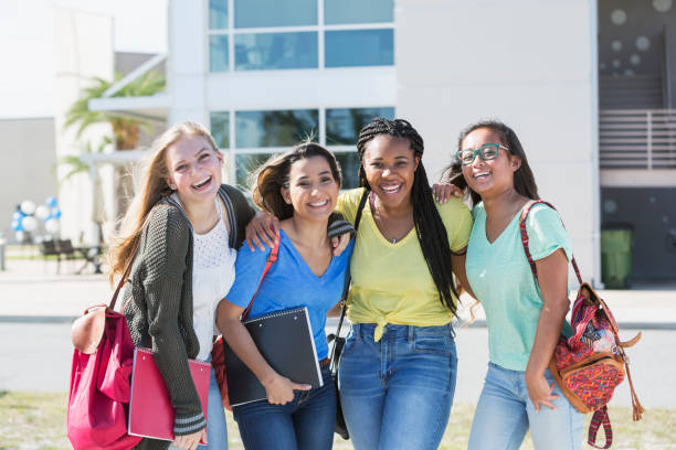 Four multi-ethnic teenage students on campus A group of four multi-ethnic teenage girls, 18 years old, standing in front of a school building carrying book bags and notebooks. They are high school seniors or freshmen in college. female high school student stock pictures, royalty-free photos & images