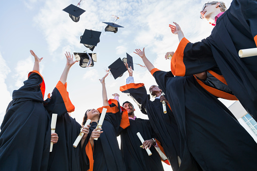 A group of seven multi-ethnic high school or university graduates wearing graduation gowns, holding diplomas. Throwing their caps in the air.
