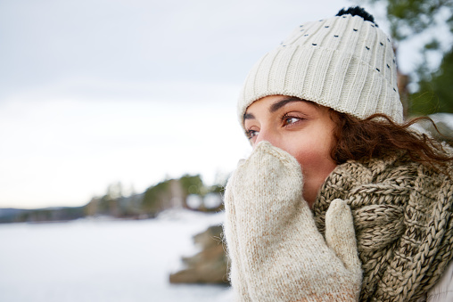 Introspective beautiful young woman warming up nose with mittens while walking in forest