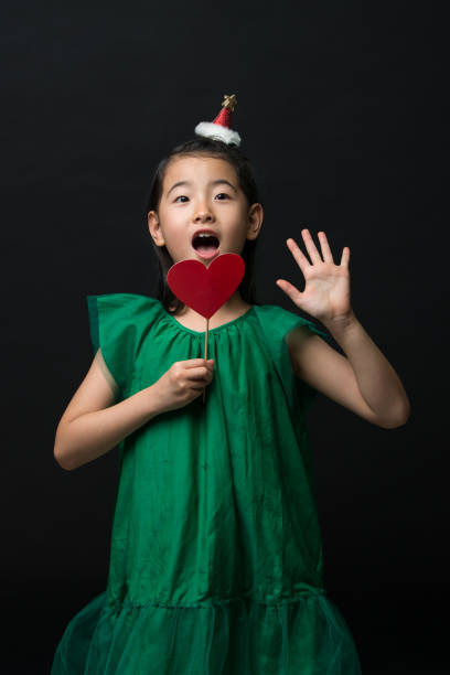 Linda Niña Asiática Vestida Con Un Vestido Verde Con Un Ornamento De La Navidad Y Un Palo De Corazón Sobre Un Fondo Negro Una Cara Sorprendida Foto de stock y más banco Linda Niña Asiática Vestida Con Un Vestido Verde Con Un Ornamento De La Navidad Y Un Palo De Corazón Sobre Un Fondo Negro Una Cara Sorprendida Foto de stock y más banco
