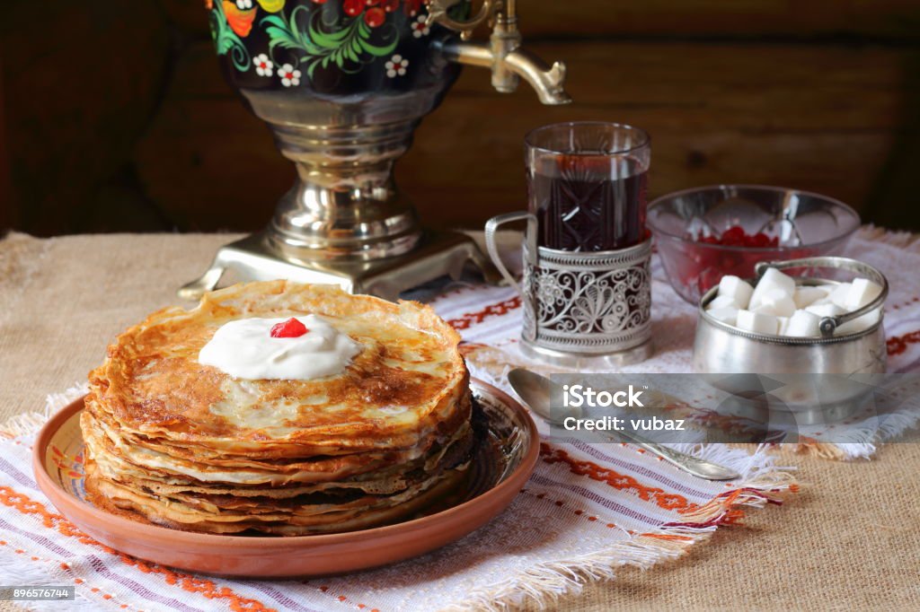 A stack of pancakes. A stack of pancakes in a clay plate and a glass of tea from a samovar on the background of a log wall. Russian Culture Stock Photo