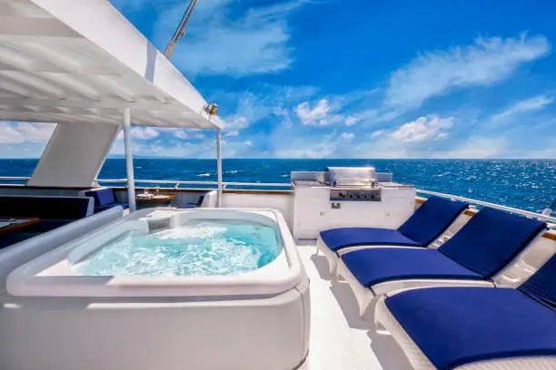 White steel yacht with white deck, nautical blue chairs, whirlpool spa with blue sky and tropical water in the background.