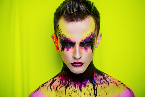 Creative man boy model purple pink yellow green colors. Bright conceptual art make-up glows light party time. Multicolored pins, clamps, clips dark hair. Artistic shiny good lifestyle. Strange emotion