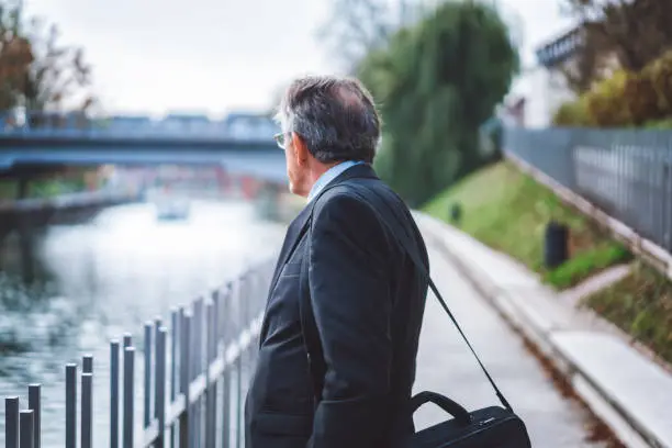 Mature and successful businessman taking a break from his work, walking next to the river, walking across the bridge, wearing a nice dark blue suit, a tie and a white shirt, wearing his glasses, having his laptop bag on shoulder, making business, feeling happy, sun is shining, walking next to the fence in the park, feeling successful and confident.