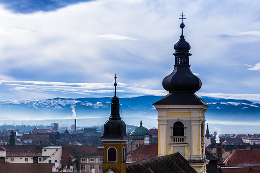 Horizontal color image depicting the ancient, medieval skyline of Sibiu, a city in the Transylvania region of Romania. In the foreground we can see the spires of old orthodox churches, and the traditional Germanic architecture of the town's buildings and houses spread out beyond. In the distance the snow-capped Carpathian mountains provide a beautiful backdrop on this winter Decemver day. Room for copy space.