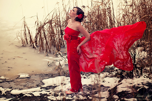Young girl in red dress stands on the coast of lake bare foot on the snow. Wind blows her dress up.