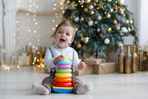 A little boy sits on a white glossy floor near a smart, green, Christmas tree with gift boxes under it, against the background of festive decorations and lights, one collects a multi-colored plastic pyramid