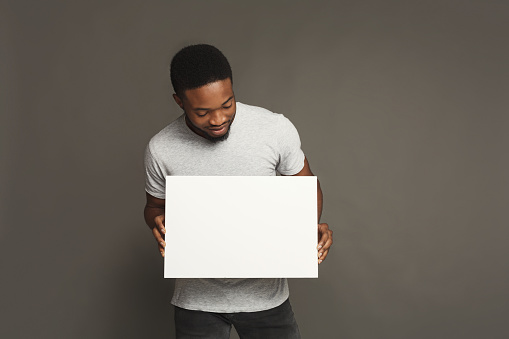 Picture of young smiling african-american man holding white blank board on grey background, copy space