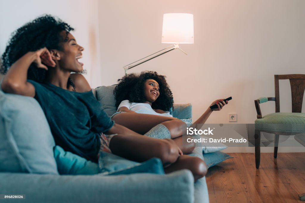 Happy young two black women lying down in the couch watching tv  "n Watching TV Stock Photo