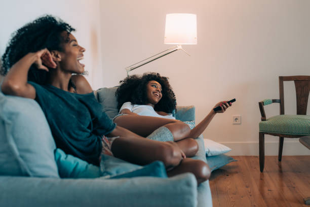 Happy young two black women lying down in the couch watching tv  "n stock photo