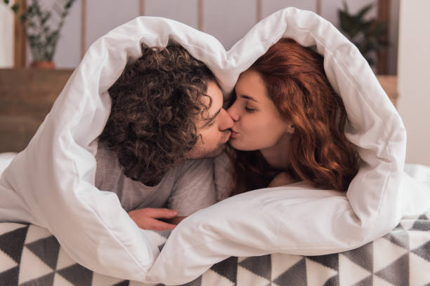 Couple at home Beautiful couple is kissing while lying in bed wrapped in blanket kissing stock pictures, royalty-free photos & images