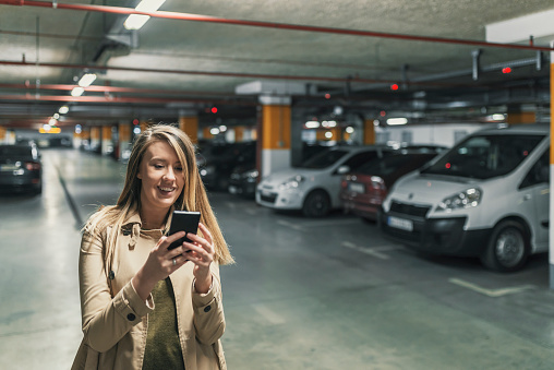 Photo of smiling Young business woman wearing beige jacket calling mobile phone while standing with blurred car in parking lot. Beautiful Caucasian female using smartphone device in underground parking with cars.