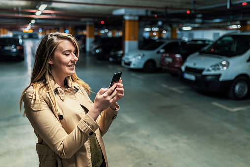 Photo of smiling Young business woman wearing beige jacket calling mobile phone while standing with blurred car in parking lot. Beautiful Caucasian female using smartphone device in underground parking with cars.