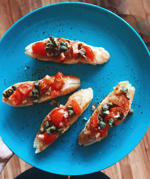 bruschetta - an italian snack with tomatoes, capers, garlic, baguette and cheese on a blue plate in the girl hand - bruschetta cutting board italy olive oil imagens e fotografias de stock