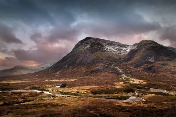 A small house nestles in the valley beneath a snow capped mountain in Glencoe. A river runs through the scene. It is wild and remote