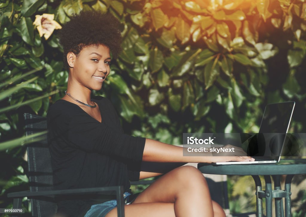 Brazilian woman in public park with net-book Smiling young African American female student sitting at the table in park and looking at camera while using her laptop; black cheerful girl with curly Afro hair working on her netbook in a garden Adult Stock Photo