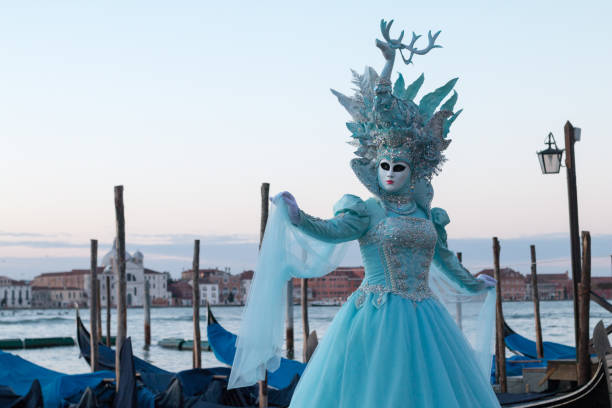 Venice, Veneto, italy - 6 february 2016: A person dressed in an elaborate costume plays a blue violin in Venice, Veneto, Italy. The outfit features a teal and gold coat with matching top hat adorned with intricate embellishments. They stand against a rustic red brick wall with a wrought iron window, characteristic of Venetian architecture. The scene evokes the vibrant atmosphere of Venice's cultural and artistic heritage, often seen during events like Carnival.