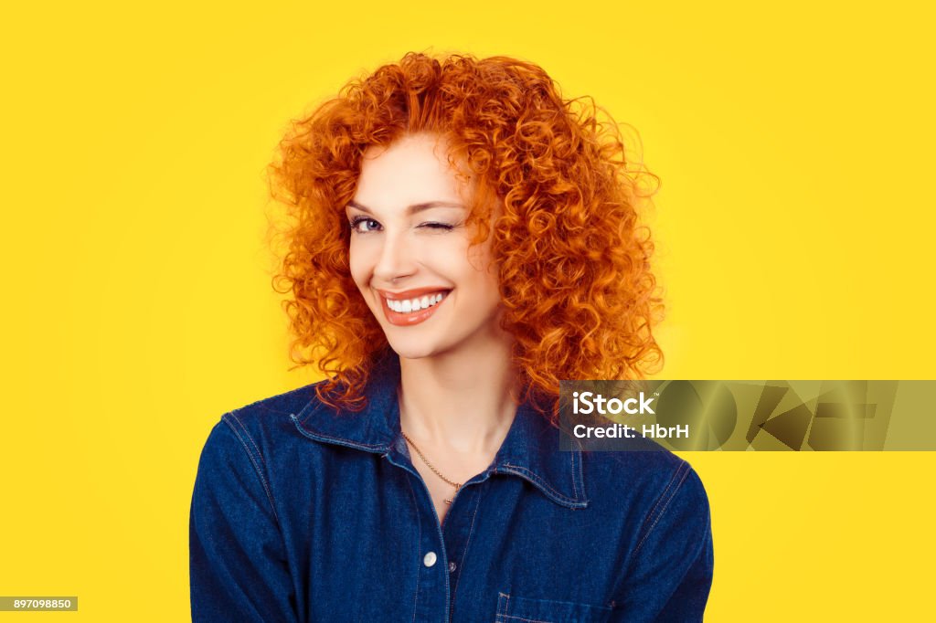 Winking. Closeup woman redhead curly hair smiling blinking eye to you camera isolated on yellow background. Happy life pictures, happiness Women Stock Photo