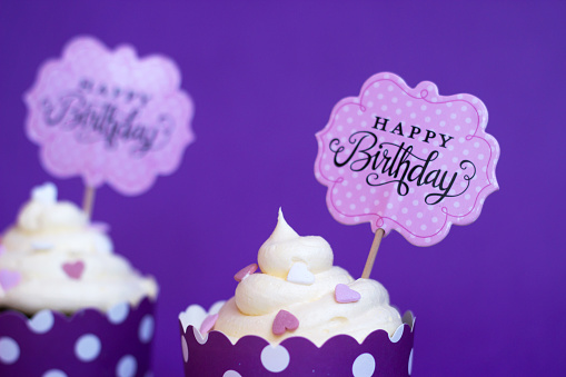 Vanilla cupcakes with small decorative hearts and happy birthday sign, against violet background; party background