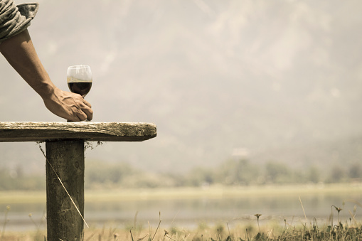 Over a wood bench, Male muscled hand holding a fine glass cup serving with red chilean wine near to a generic wine glass bottle. On back, a landscape view of lake shore in Chilean Patagonia with Los Andes mountain range near to Torres del Paine National Park and Bariloche, Argentina in a afternoon day. Chilean wine is one of the most famous wines around the world, as Australia, France, California, Italy as well. Open Shoot landscape with a copy space.