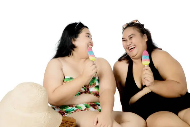 Summer concept. Two obese women wearing bikini while eating ice cream and laughing together, isolated on white background