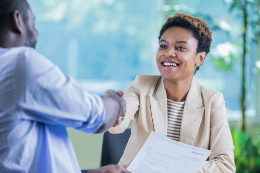 Confident young businesswoman greets a male job candidate. They are shaking hands. The woman is holding the man's resume.