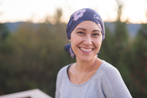 A beautiful young woman wearing a head wrap looks toward the camera and smiles radiantly. She is standing outdoors and there are mountains and trees in the background.