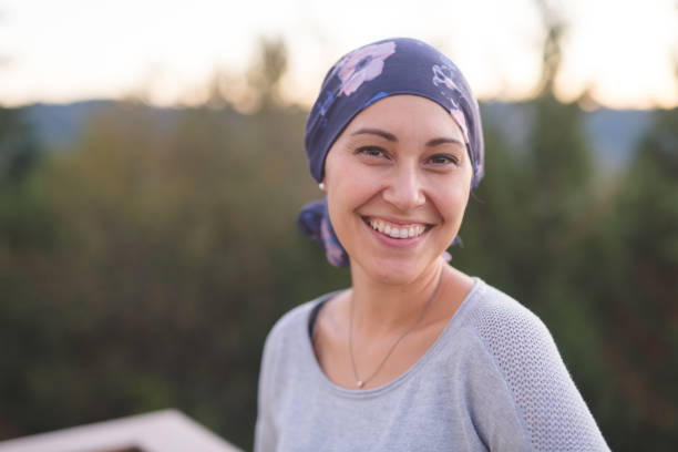 Beautiful Woman with Cancer Smiles A beautiful young woman wearing a head wrap looks toward the camera and smiles radiantly. She is standing outdoors and there are mountains and trees in the background. brest cancer hope stock pictures, royalty-free photos & images