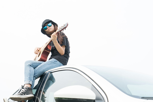 style girl sitting on car roof playing guitar against sky.