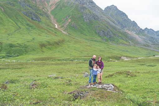Two parents and their two children take a break from hiking to pose for a picture by an Alaskan mountain. Mom is kissing dad on the cheek. They are embracing each other...but the kids look like they are done with pictures!