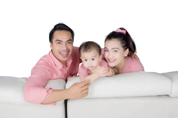 Young parents with their baby smiling at the camera while sitting on the couch, isolated on white background