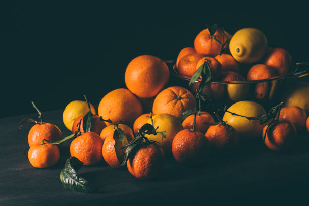 close up view of pile of lemons and tangerines with leaves in strainer on dark surface