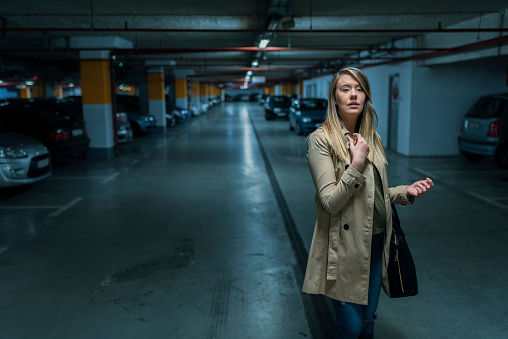 Photo of Serious attractive, Young business woman wearing beige jacket, standing with blurred car in parking lot. Beautiful Caucasian female  in underground parking with cars.  Brown hair Woman is walking dangerously in a car parking area while looking for her car at night. Girl in the deep thinking about her next destination while looking away.
