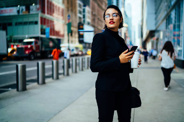istock Trendily dressed female manager holding smartphone checking mail while strolling on manhattan street,beautiful businesswoman with red lips and in eyeglasses walking with coffee to go sending messages 897391274