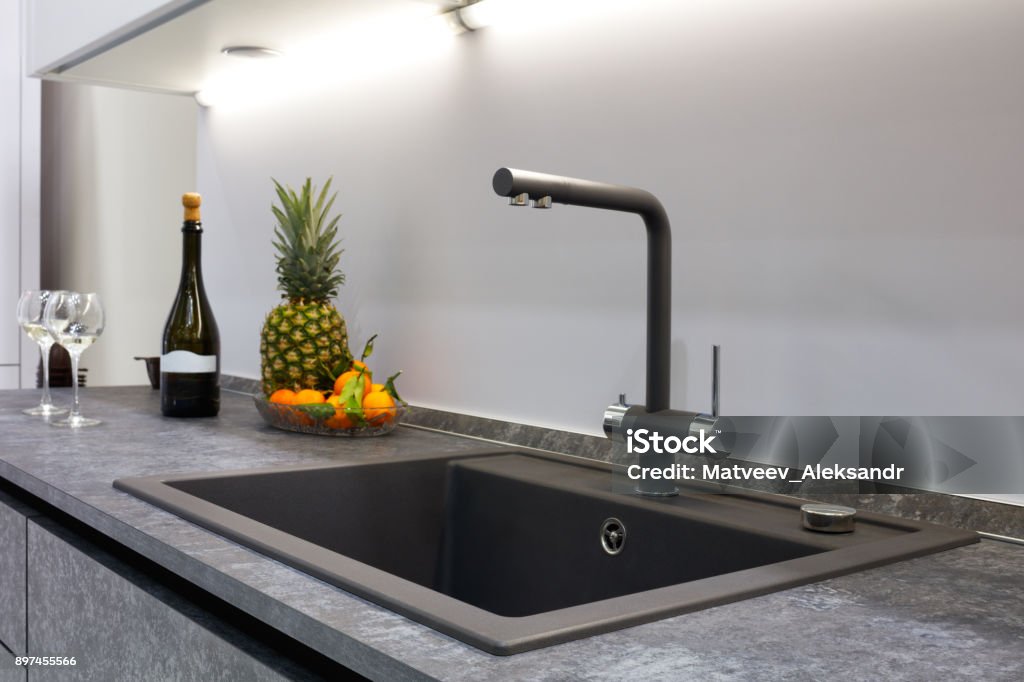 The interior of the modern kitchen is illuminated with a gray stone countertop with a luxury washbasin and mixer faucet, fruit pineapple and tangerines, a bottle with red wine and two glasses. The interior of the modern kitchen is illuminated with a gray stone countertop with a luxury washbasin and mixer faucet , fruit pineapple and tangerines, a bottle with red wine and two glasses. Bar Counter Stock Photo