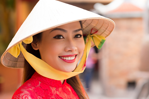 Head and shoulders portrait of attractive Vietnamese woman wearing conical hat looking away with charming smile, blurred background