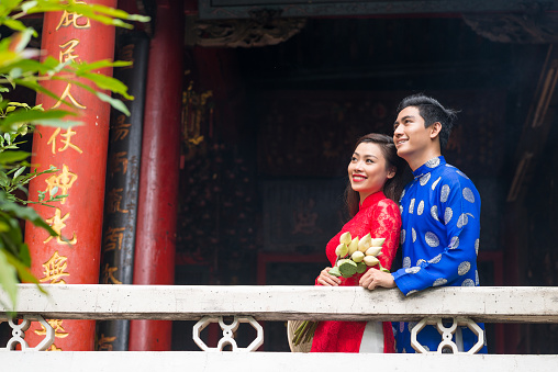 Loving Asian couple wearing traditional costumes looking away with toothy smiles while enjoying picturesque view from terrace decorated with characters