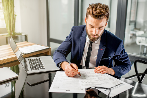 Handsome tax manager dressed in the suit working with documents and laptop at the modern office interior