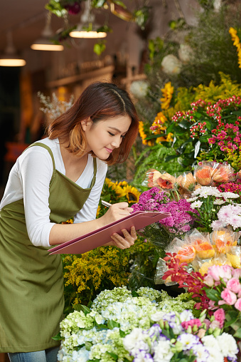 Attractive Asian flower shop owner wearing apron writing down necessary information while taking inventory, portrait shop