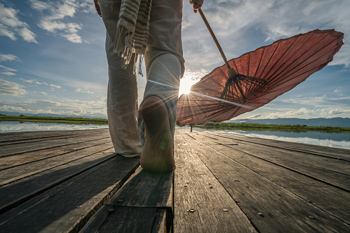 Woman standing on wooden pier above lake at sunrise, holding a traditional Burmese umbrella. People travel solo and like locals concept.