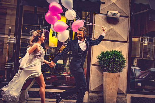 Affectionate bride and groom, holding hands, happily run down the street. Bridegroom carries a bunch of white and pink balloons which flutter behind them.