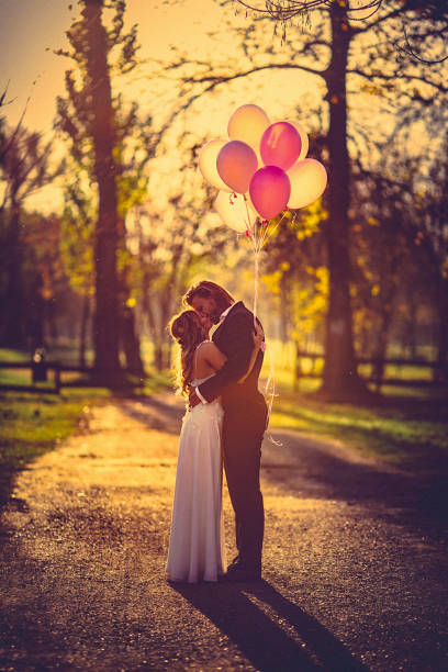 affectionate bride and groom, embraces and kisses amidst the gravel path at the park at sunset. - silhouette kissing park sunset imagens e fotografias de stock